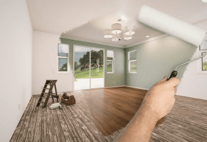 Hand uses a paint roller to reveal a renovated living room with green walls, wood floor, and patio doors.