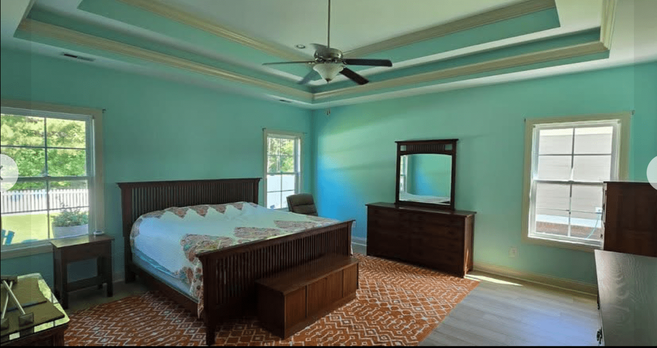 Large bedroom with teal walls, dark wood furniture, bright windows, and a ceiling fan in a recessed tray ceiling.