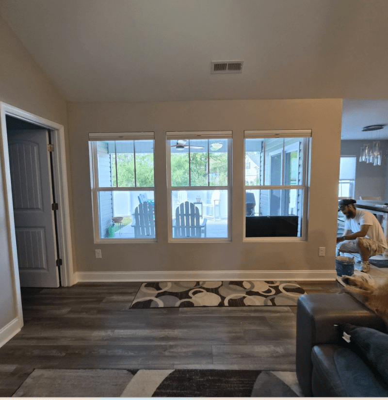Neutral living room with three windows, gray flooring, a patterned rug, and a worker painting near the kitchen.