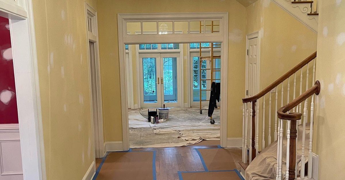 A home interior prepared for painting, with protected floors, taped surfaces, and painting supplies in a hallway.