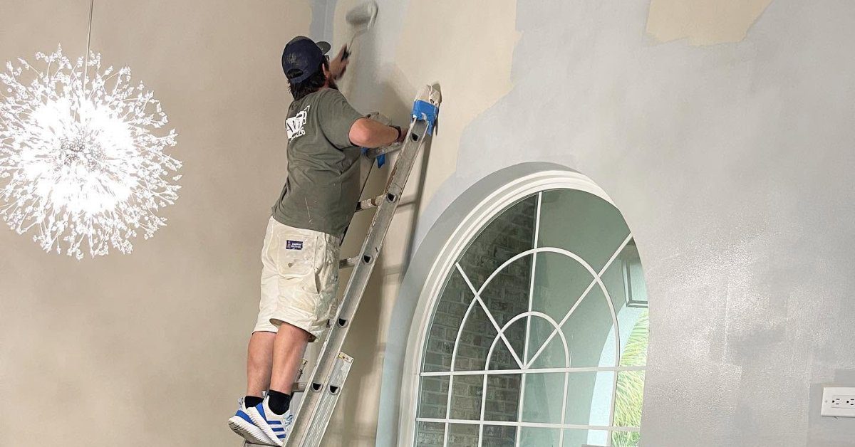 A professional painter on a ladder applying fresh paint to a high interior wall above an arched entry window.