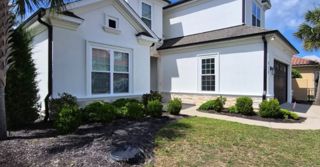 White two-story stucco home with black roof, landscaped shrubs, lawn, and a palm tree beneath a bright blue sky.