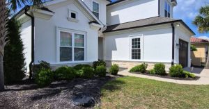 White two-story stucco home with black roof, landscaped shrubs, lawn, and a palm tree beneath a bright blue sky.