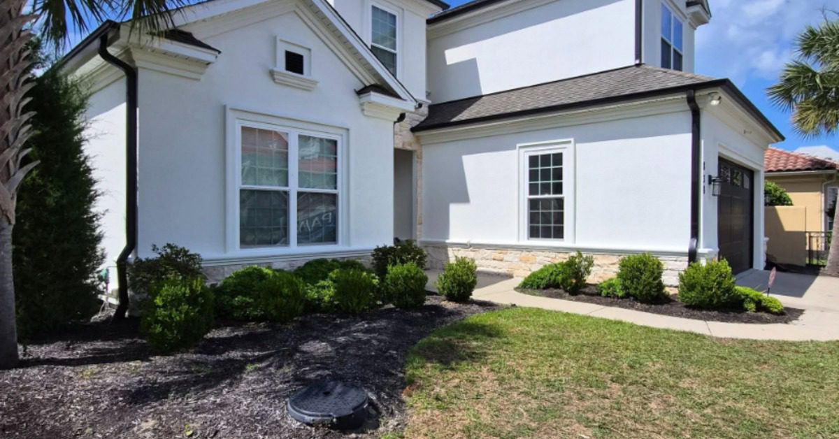 White two-story stucco home with black roof, landscaped shrubs, lawn, and a palm tree beneath a bright blue sky.