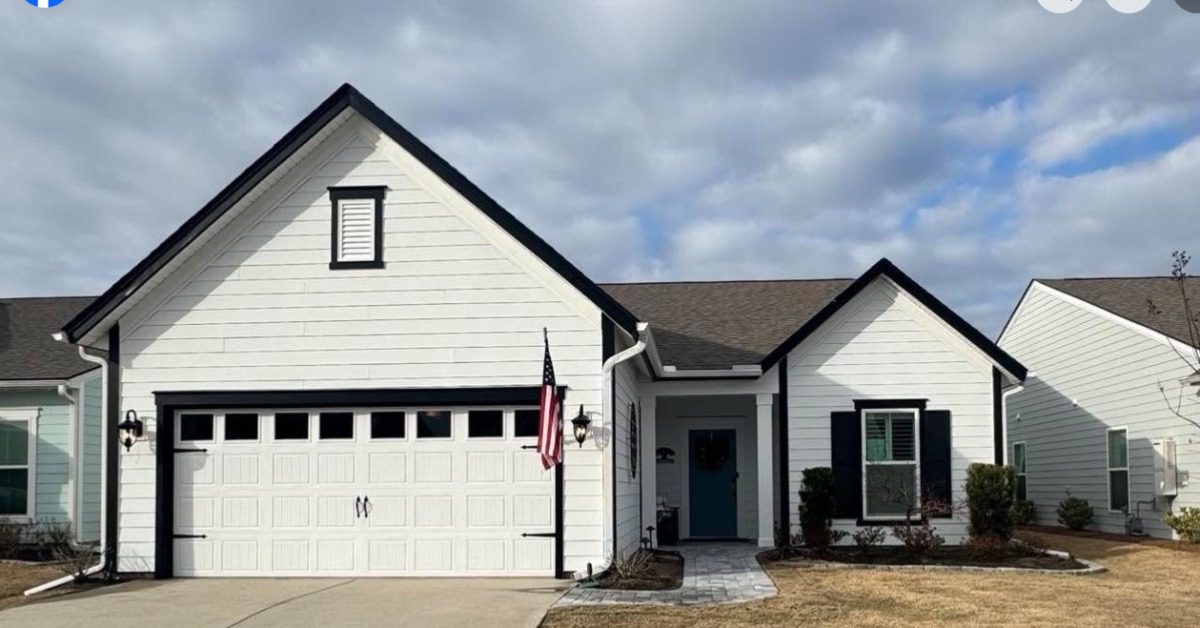 White single-story home with black trim, attached garage, blue front door, paved walkway, and cloudy sky overhead.