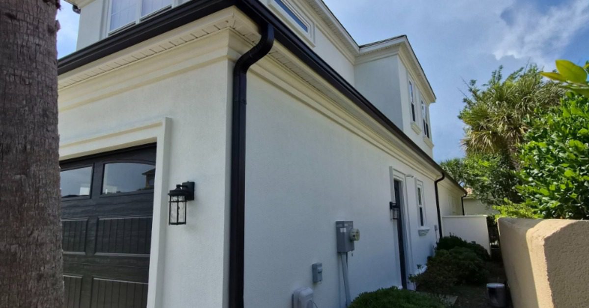 Side view of a white two-story stucco home with black garage door, black gutters, shrubs, and palm trees nearby.
