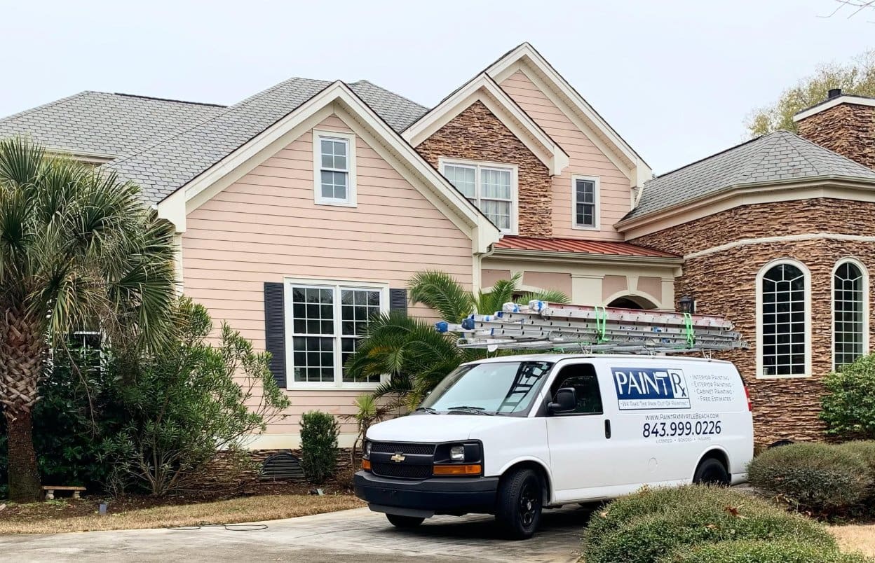 Painting service van carrying ladders is parked outside a large two-story home with pink siding and stone accents.