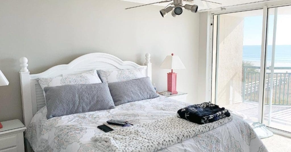 Bright bedroom with a white bed facing sliding doors that open to a balcony overlooking the ocean in daylight
