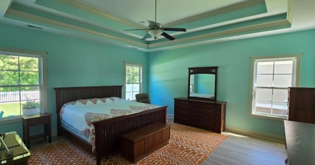 Spacious bedroom with turquoise walls, a wooden bed set, tray ceiling, and sunlight through multiple windows