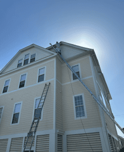 Painter works from a long extension ladder to paint upper exterior trim on a tall house under a bright blue sky.