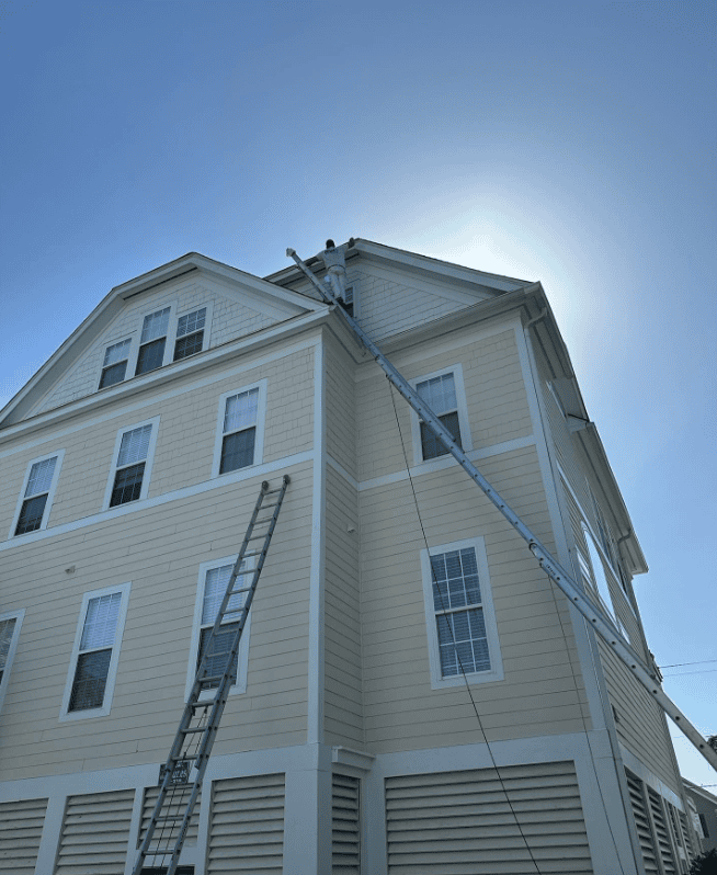 Painter works from a long extension ladder to paint upper exterior trim on a tall house under a bright blue sky.