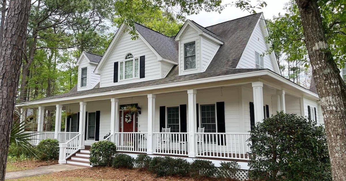White two-story house with black shutters, a wraparound porch, red front door, and tall trees surrounding it.