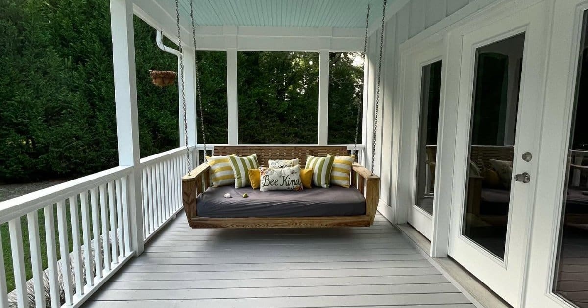 Wooden porch swing with striped pillows hangs beneath a white ceiling fan on a bright, covered home porch.