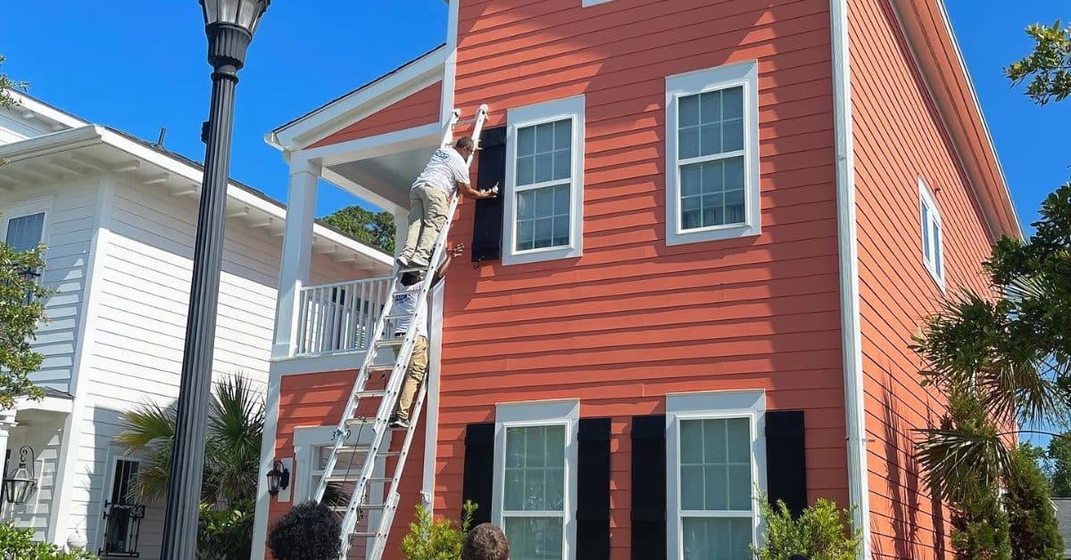 Four painters work outside a tall red house with white trim, black shutters, and a ladder reaching the upper floor.