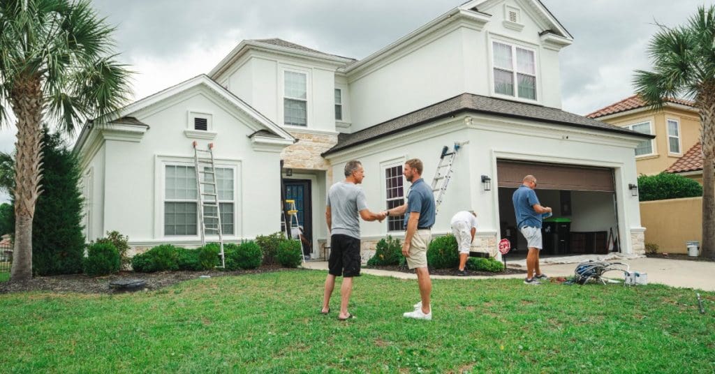 Workers gather outside a large white two-story home with ladders and equipment as two men shake hands.