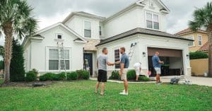 Workers gather outside a large white two-story home with ladders and equipment as two men shake hands.