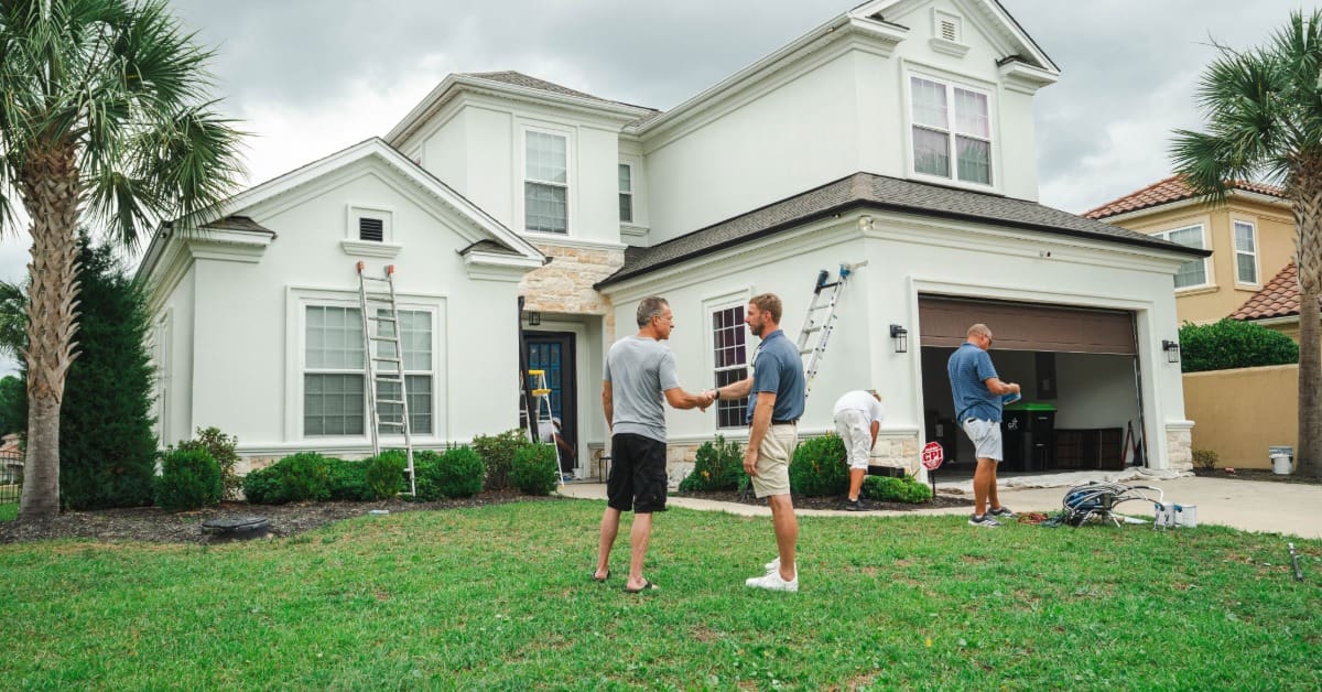 Workers gather outside a large white two-story home with ladders and equipment as two men shake hands.