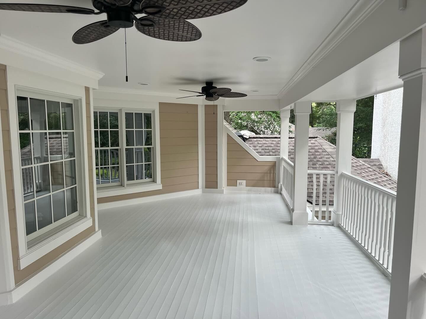 Bright covered veranda with white columns, railings, painted floors, large windows, and two dark ceiling fans.