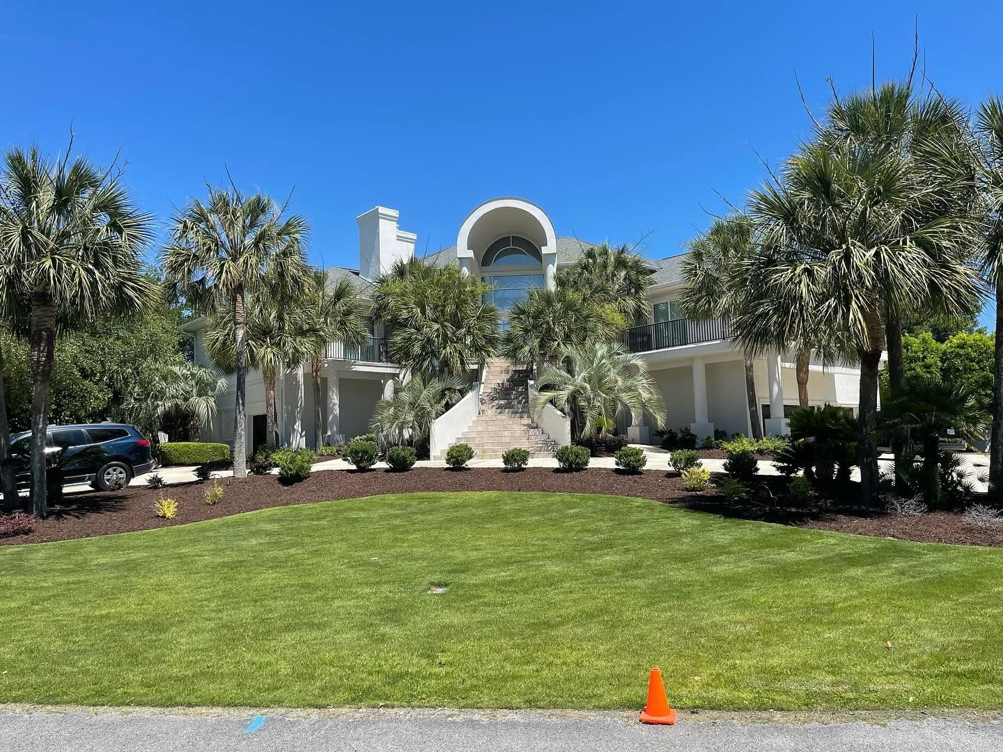 Large white two-story home framed by palm trees, with a broad lawn, central staircase, and clear blue sky.