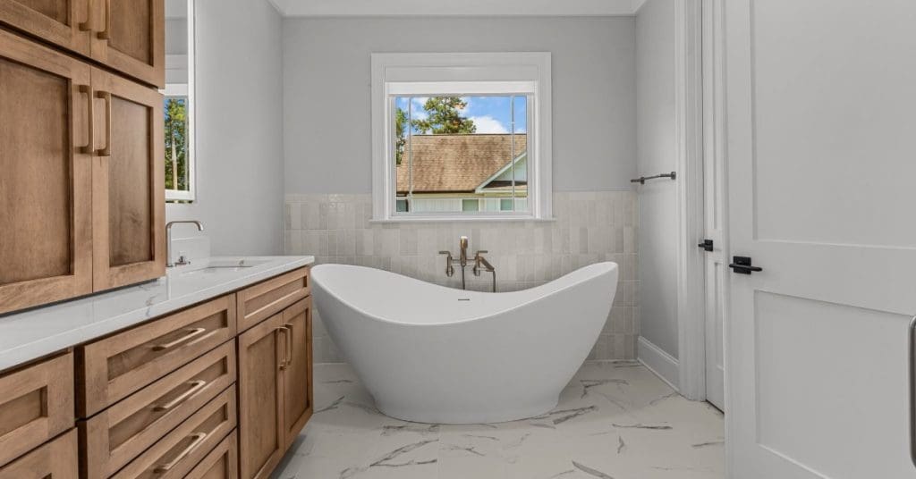 Modern bathroom with a white freestanding bathtub beneath a window, wooden vanity cabinets, and marble-look flooring.